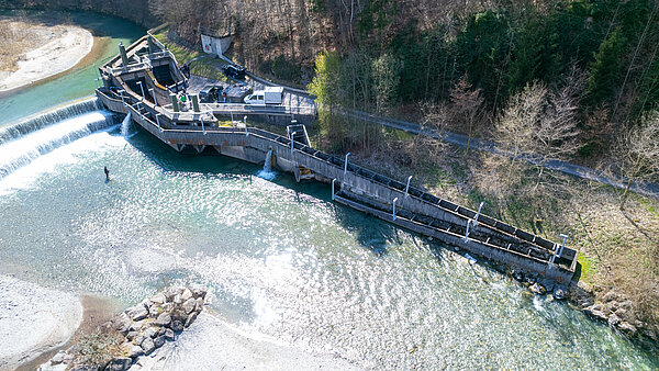 Aerial view of a hydro-electric plant and reservoir on a river with overflowing water. The system shows platforms, technical equipment and fall protection systems for maintenance.