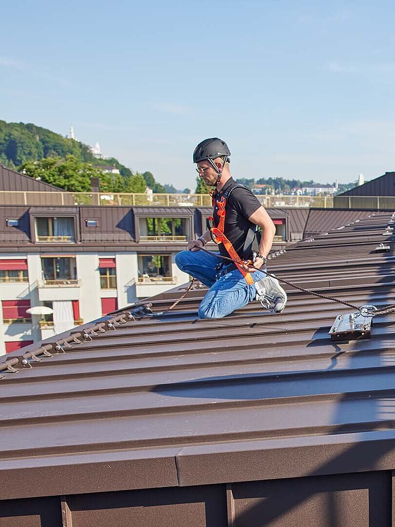 Two men are kneeling on a flat roof, wearing anti-fall PPE on their bodies.