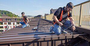 Two men are kneeling on a flat roof, wearing anti-fall PPE on their bodies.